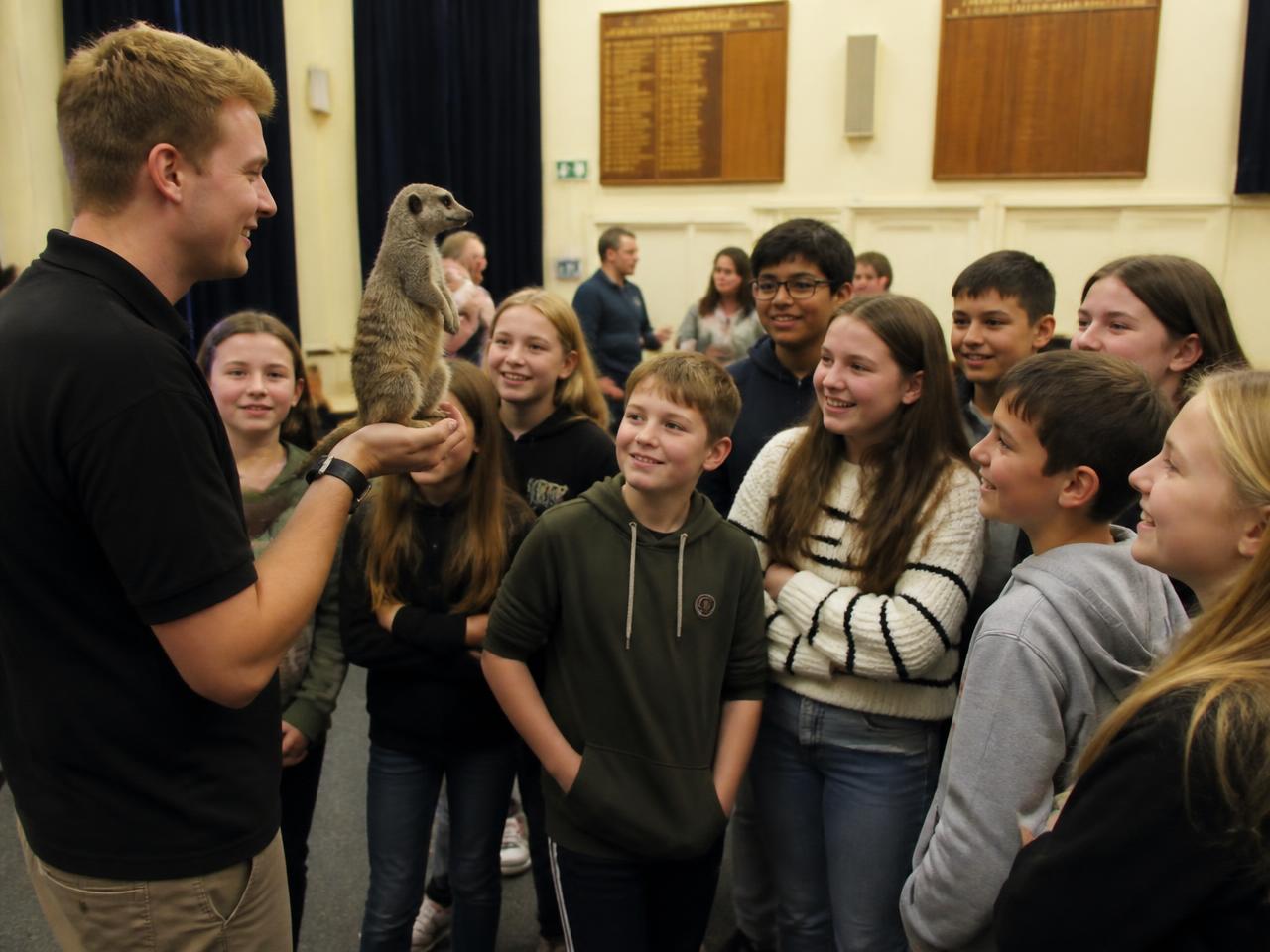 A show presenter holding a meerkat while a group of young people watch closely in a community hall